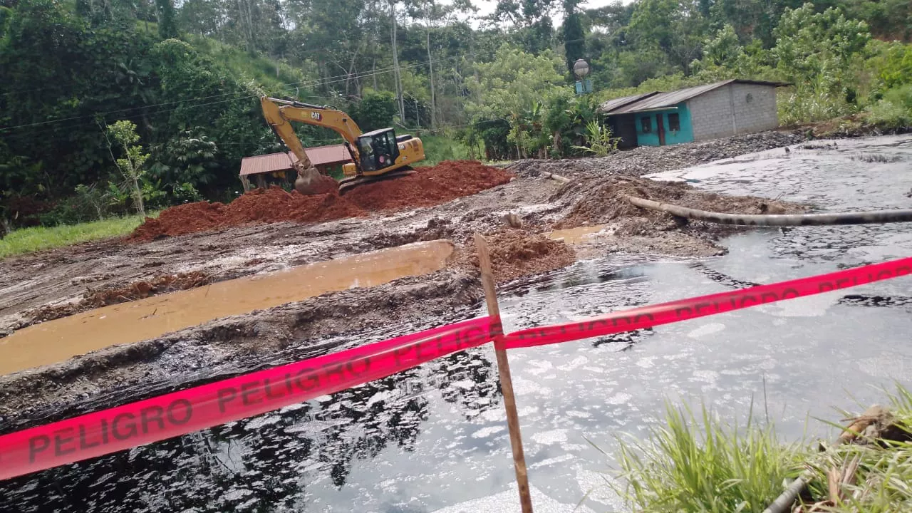 Piscina de desechos petroleros con cinta de peligro y una excavadora removiendo tierra junto a una casa